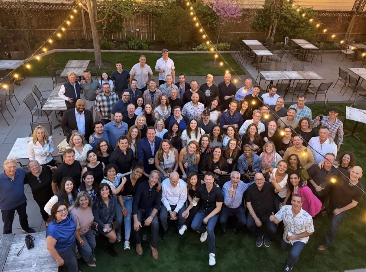 The reunited news team, several rows deep, under string lights in a beer garden at golden hour.