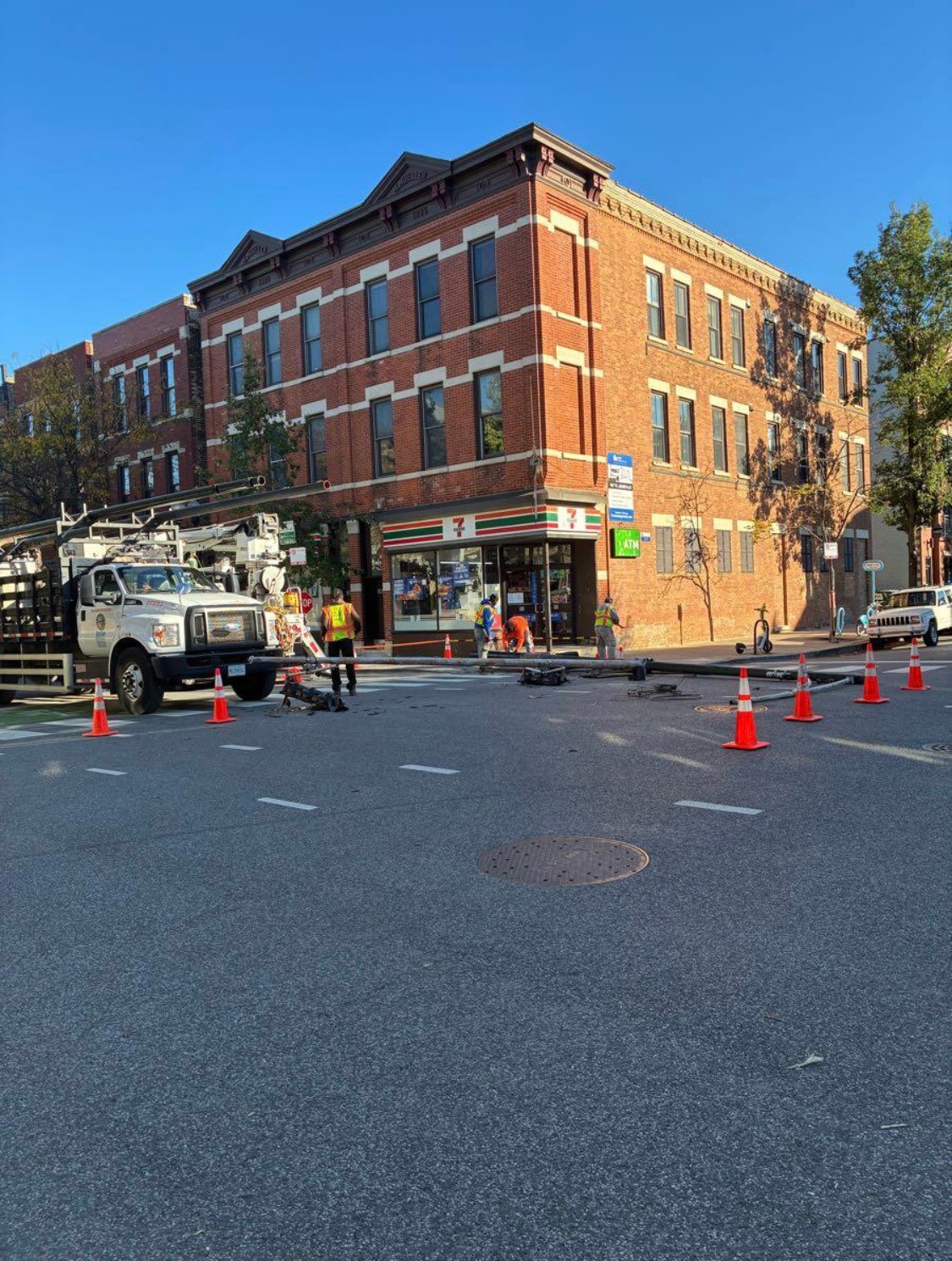 Workers, a truck and traffic cones beside fallen equipment at a red-brick corner building